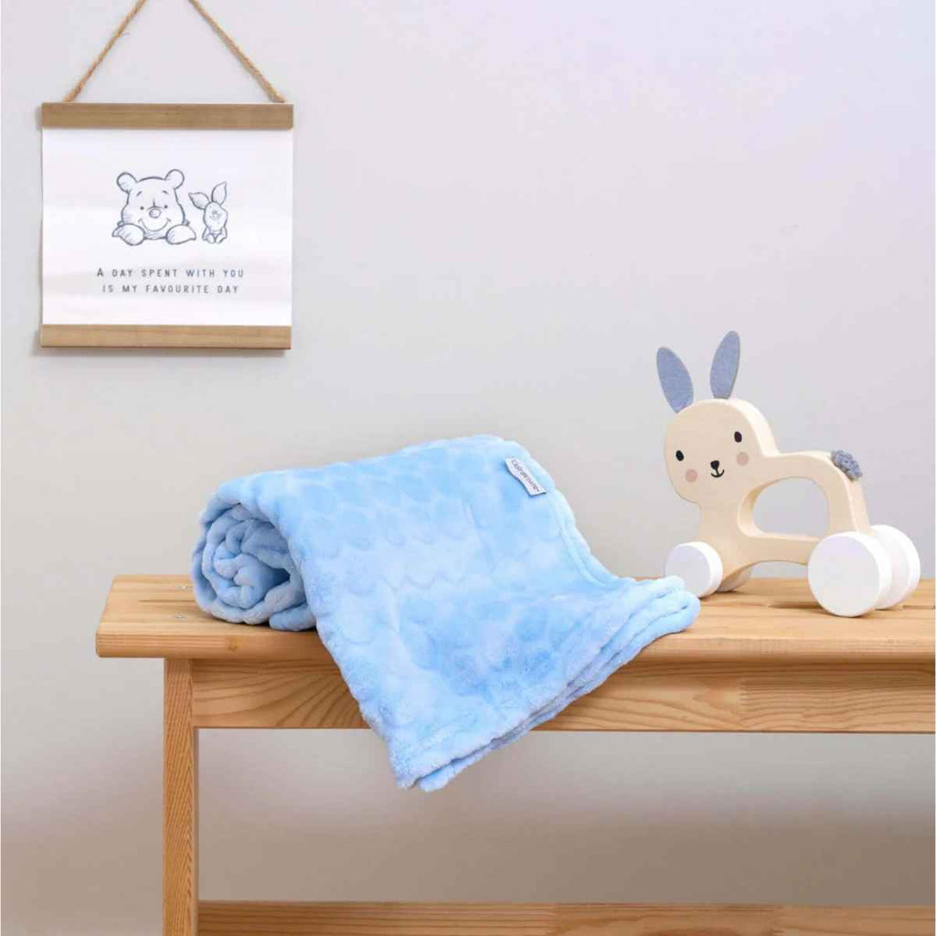 Blue baby blanket and wooden toy on a wooden surface with a framed picture in the background.