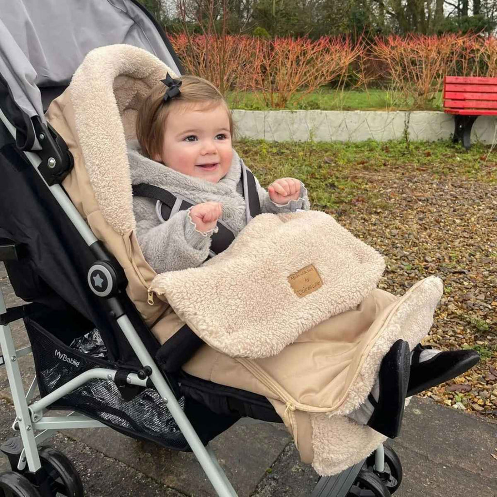 Child in a stroller with a beige footmuff, sitting outdoors on a path.