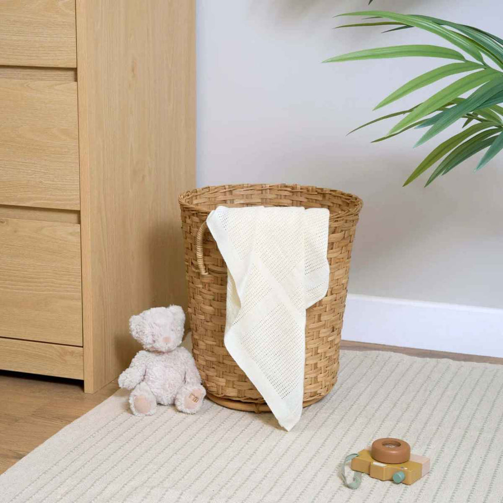 Wicker laundry basket with cream blanket over the top and teddy bear on the floor.