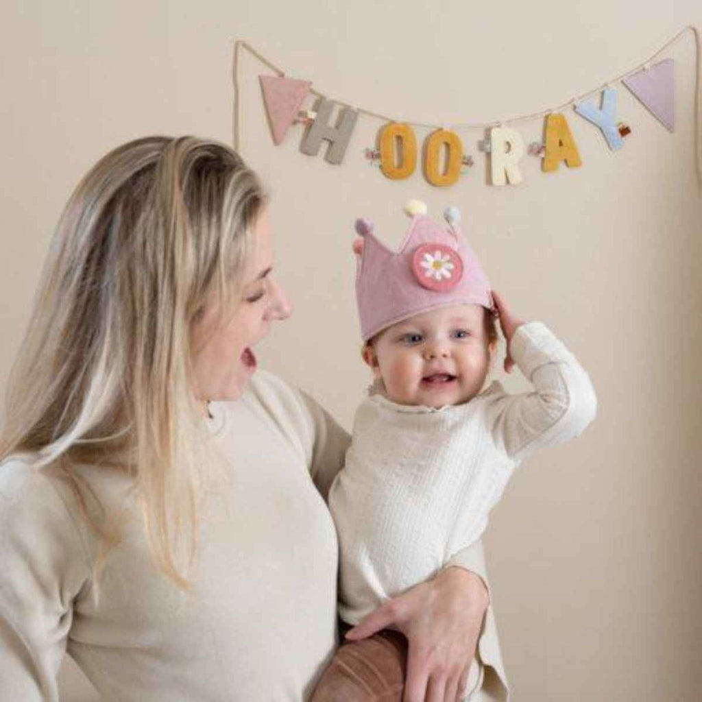 Woman holding a baby wearing a pink birthday crown with a 'HOORAY' banner in the background