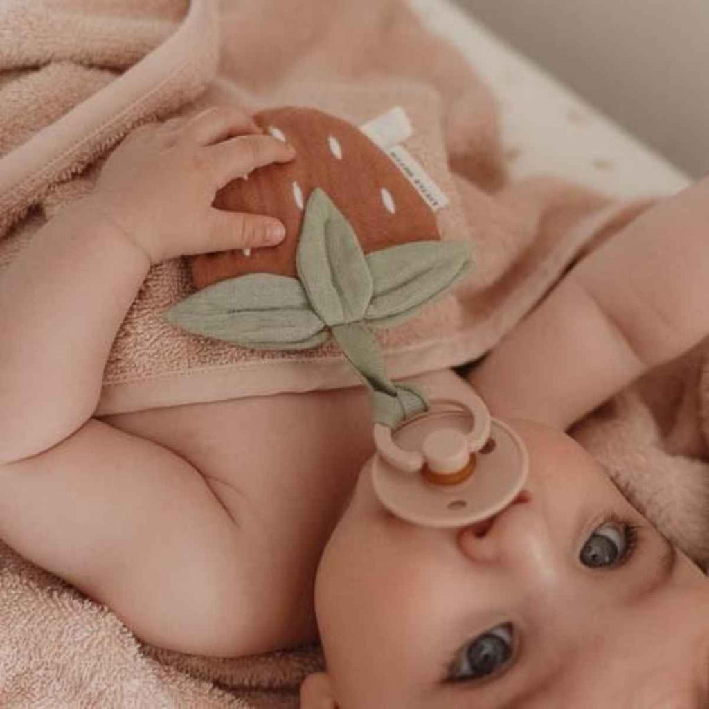 Baby holding strawberry pacifier cloth with a pacifier, lying on a soft surface.