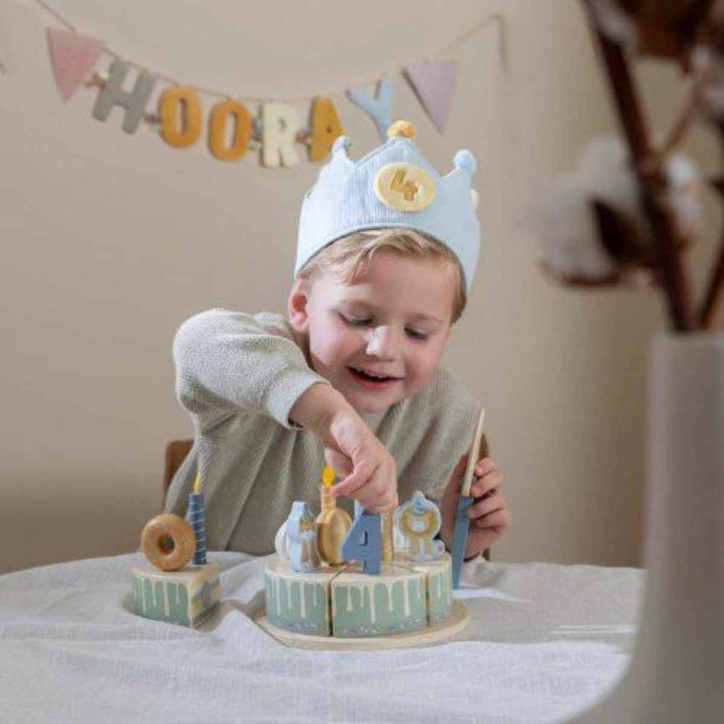 Child wearing a blue birthday crown playing with a toy cake set in a room with 'Hooray' decorations.
