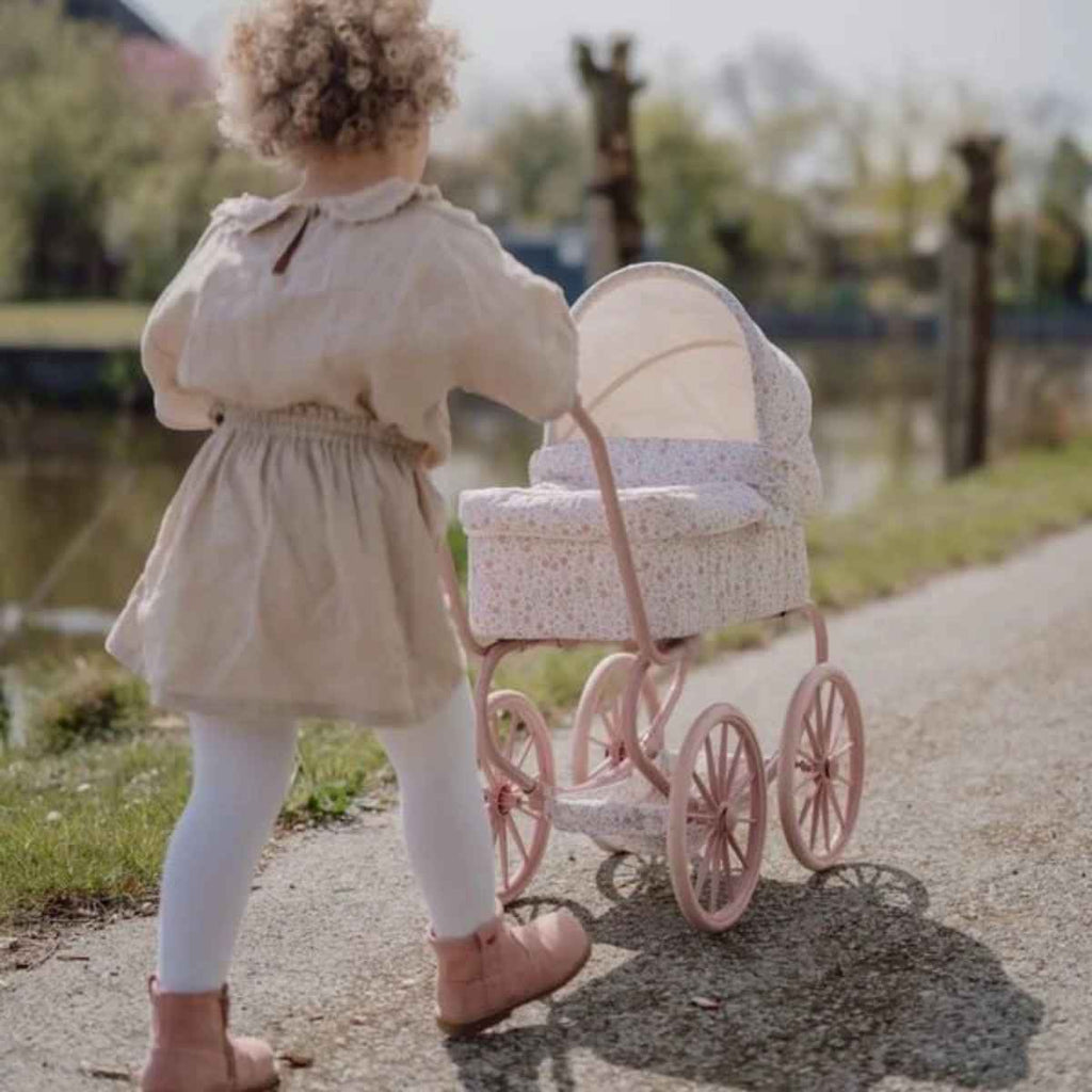 Child in a beige dress pushing a pink doll pram outdoors.