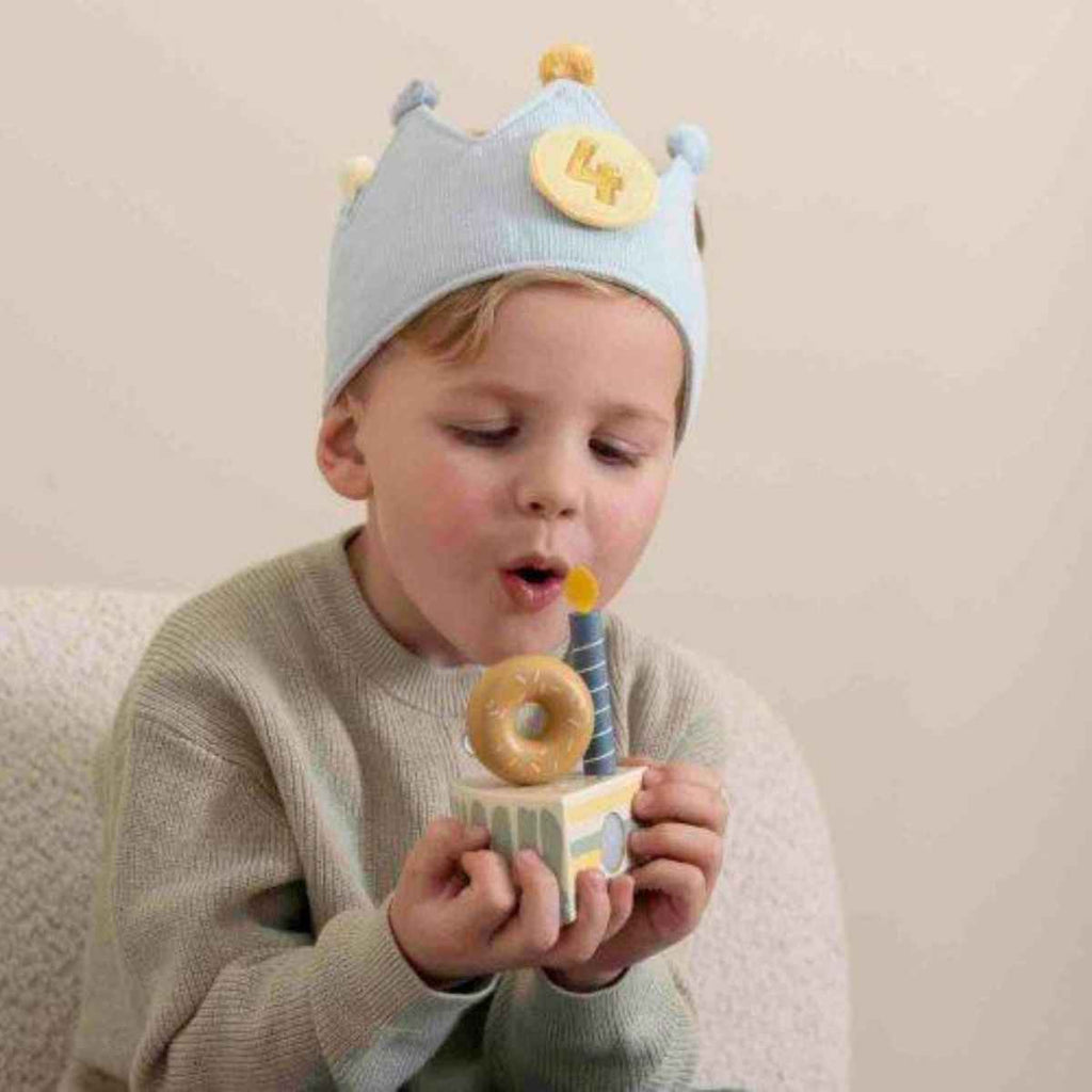 Child wearing a blue birthday crown with a number '4' and holding a wooden toy cake blowing out a pretend candle.