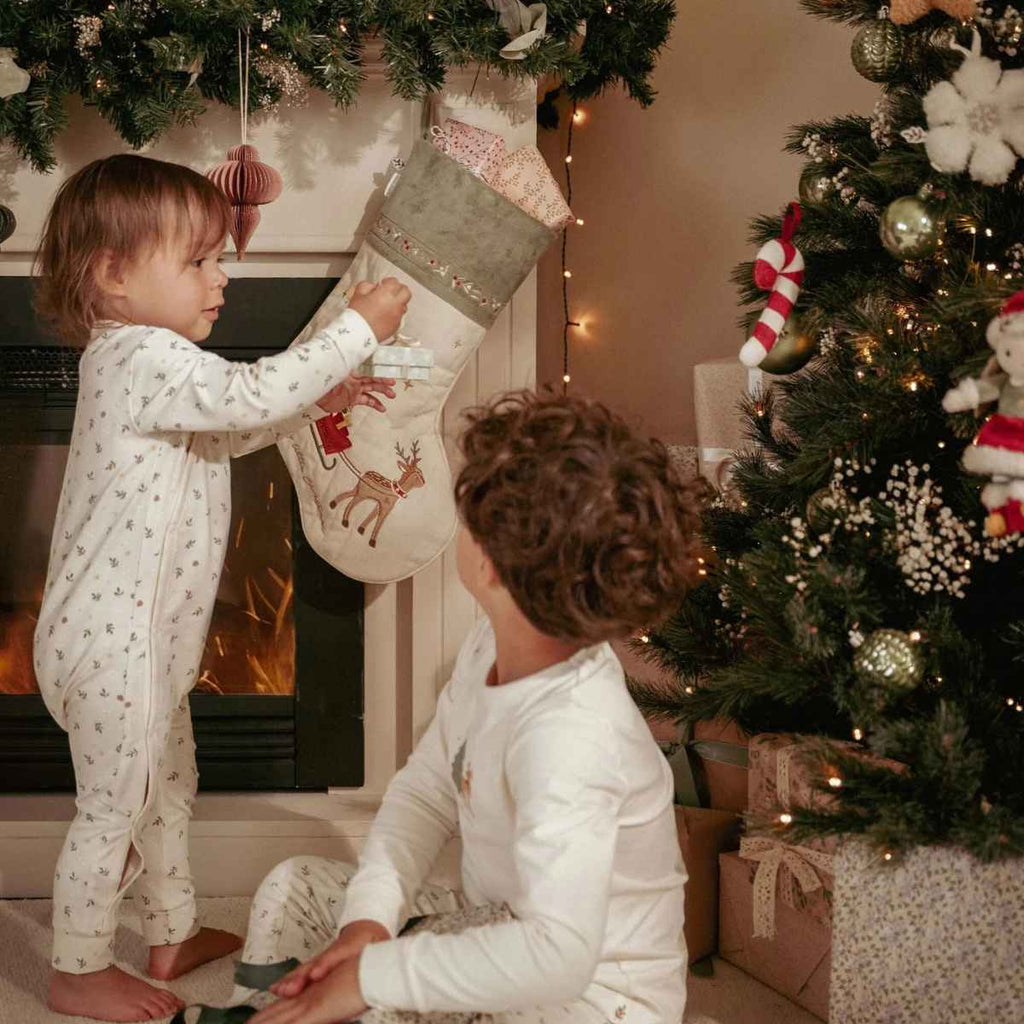 Two children in pyjamas by a Christmas tree and fireplace with a stocking hanging from it.