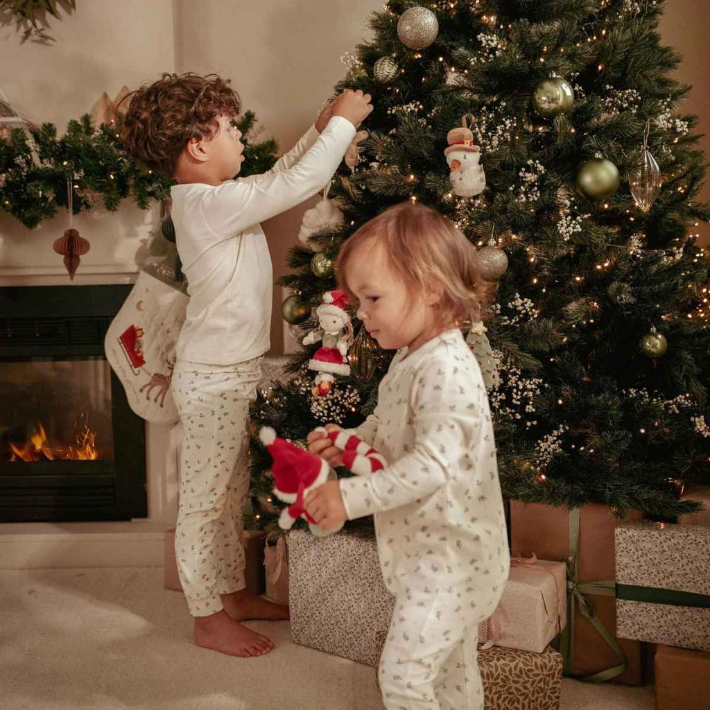 Two children in pajamas decorating a Christmas tree in a cozy living room with a fireplace.