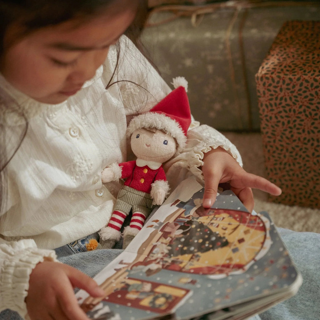 Child holding a Christmas book and a elf doll wearing a Santa hat.
