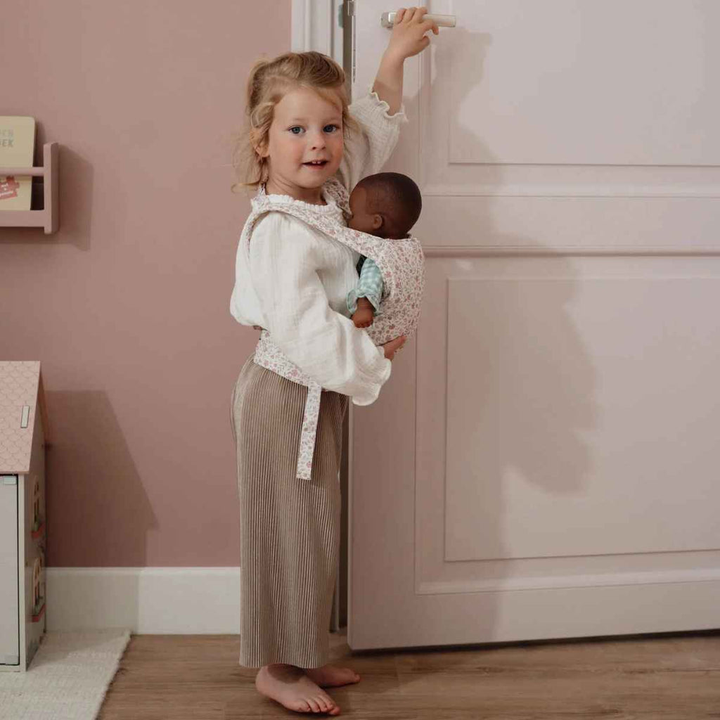 Child holding a baby doll in a carrier in a room with pink walls and wooden floor.