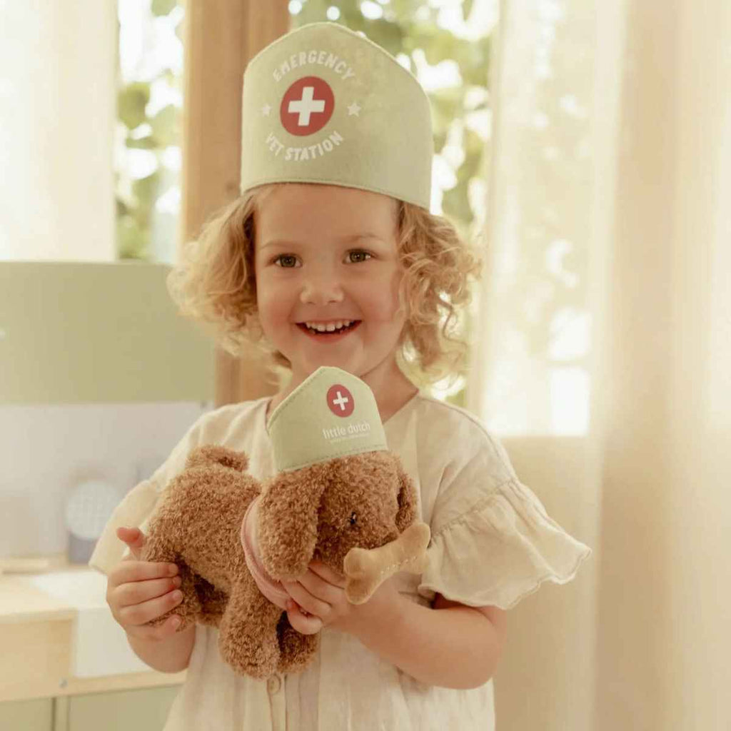 Child wearing a vet's hat with a teddy dog, smiling in a bright room.