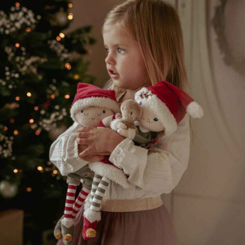 Child holding two stuffed toys with Santa hats in front of a decorated Christmas tree.
