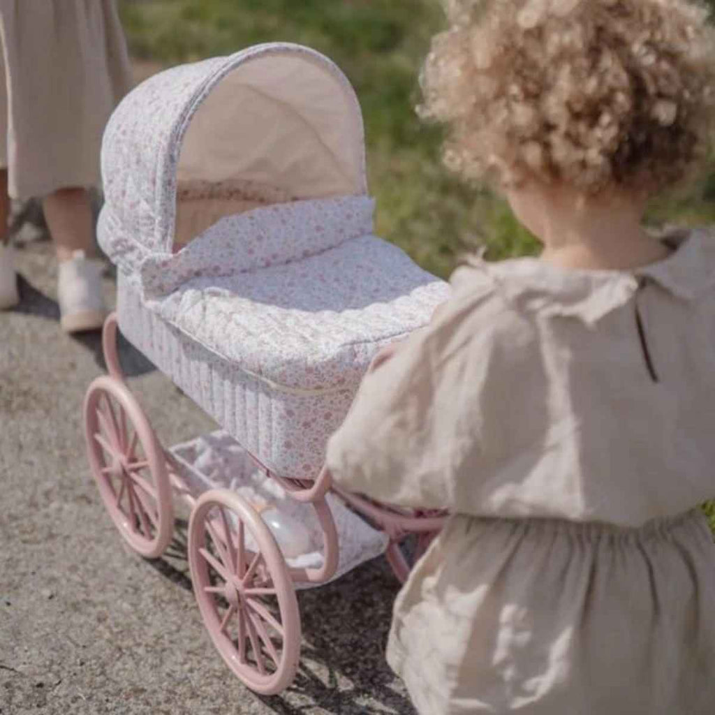 Child in a white dress standing next to a vintage-style baby buggy with pink wheels.
