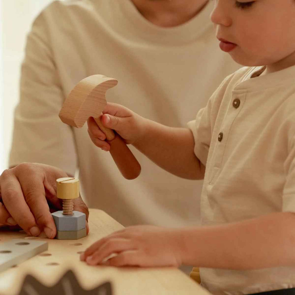 Child playing with wooden tools on a table, with an adult partially visible.