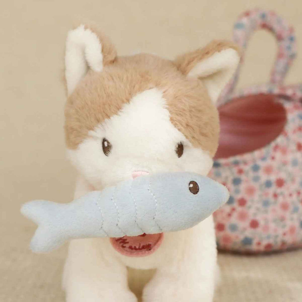 Plush toy cat holding a fish with a floral-patterned bag in the background