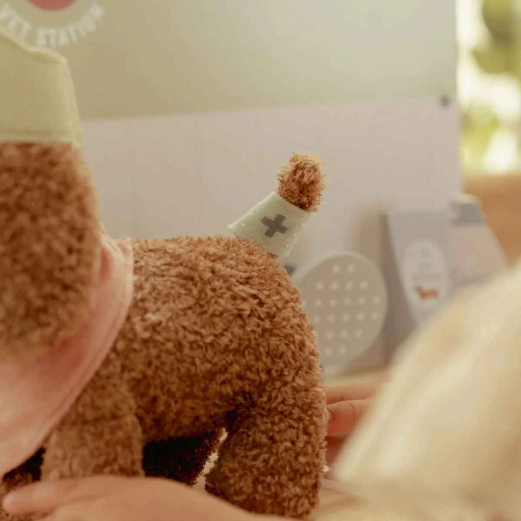 Close-up of a brown dog plush toy with a bandage on tail held by a child in a room with soft lighting.