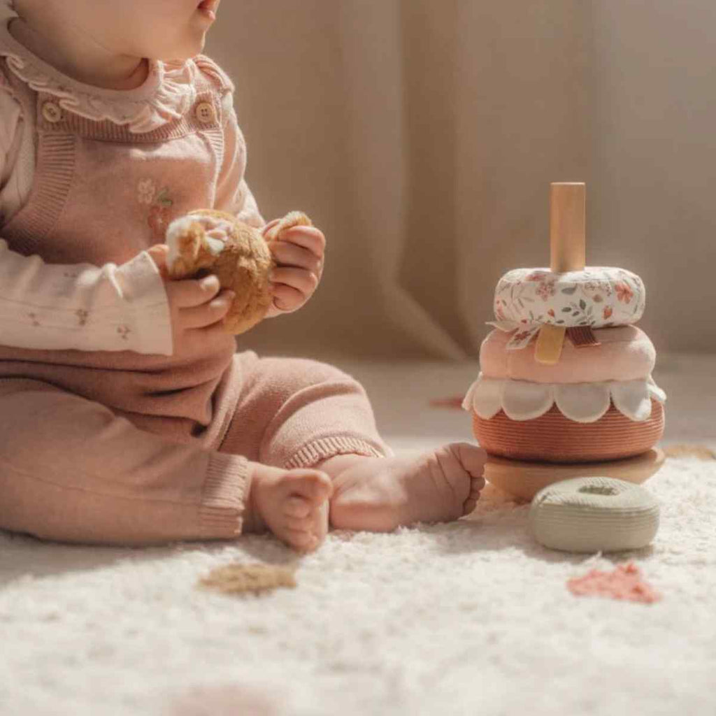 Child in pink outfit holding a teddy deer with a stack of soft stacking toys on a soft surface