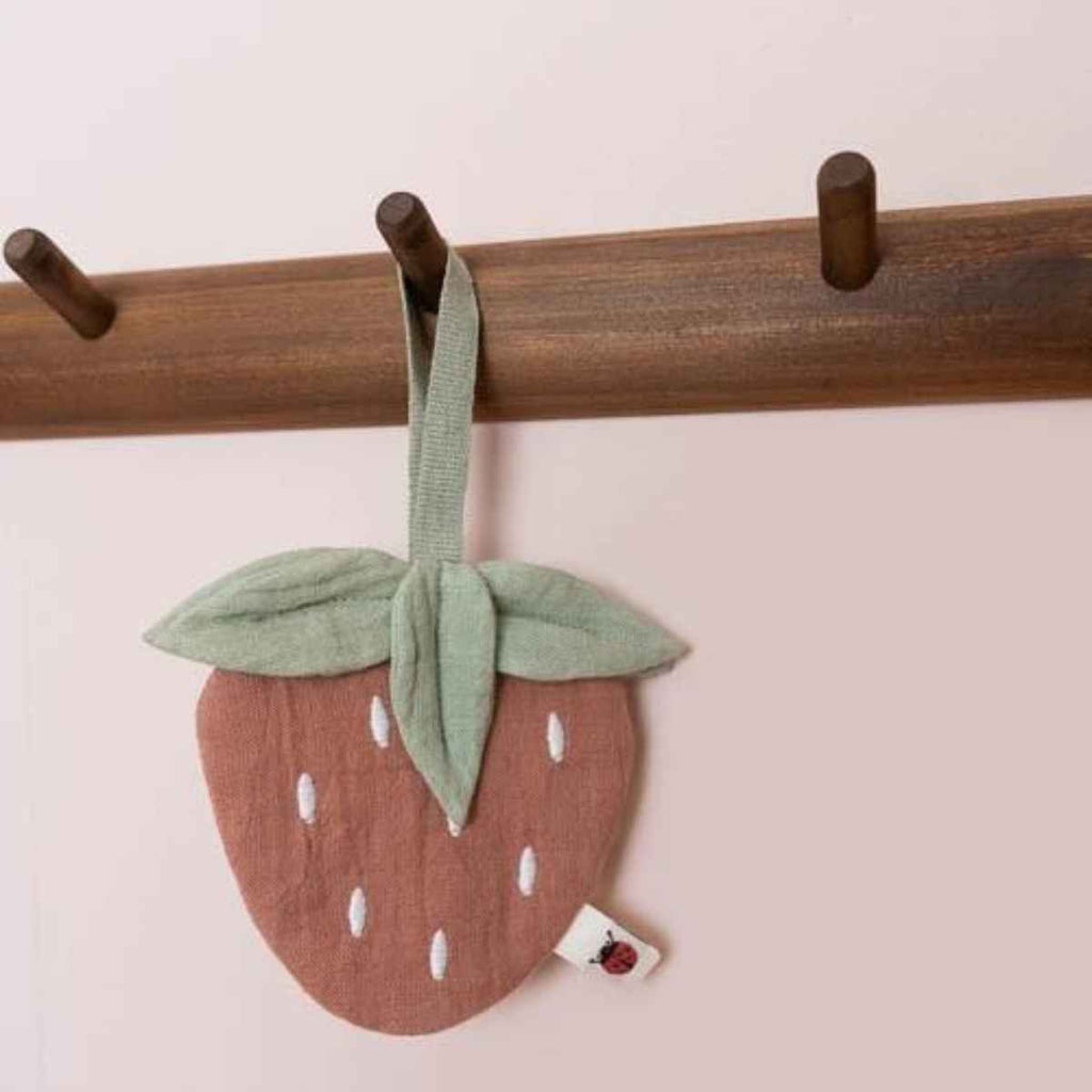 Strawberry-shaped Pacifier cloth hanging on a wooden rack against a light pink background